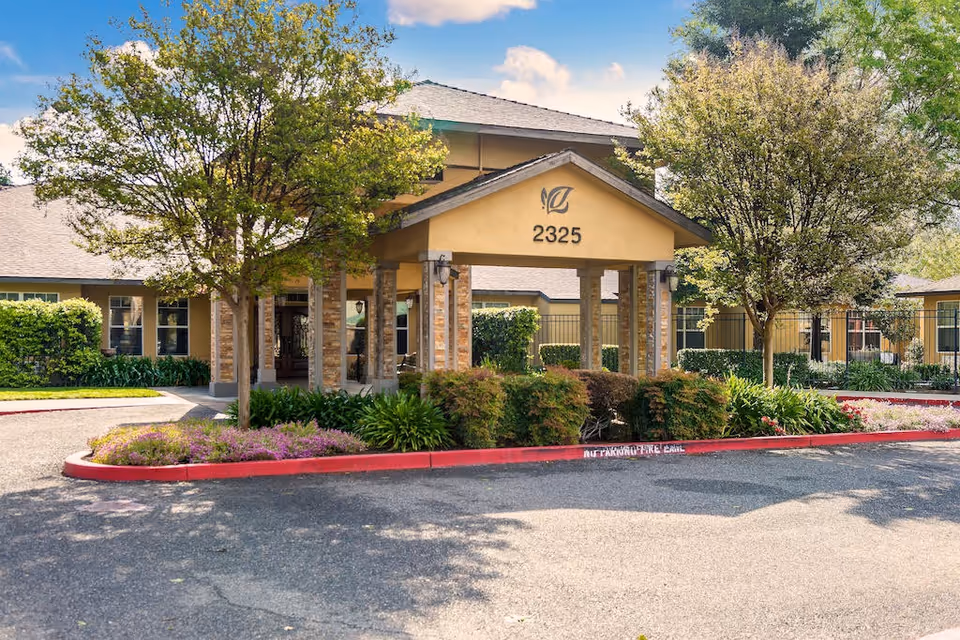 Exterior view of The Gardens of Modesto senior living facility entrance with a covered driveway, surrounded by trees, bushes, and flowers under a partly cloudy sky.