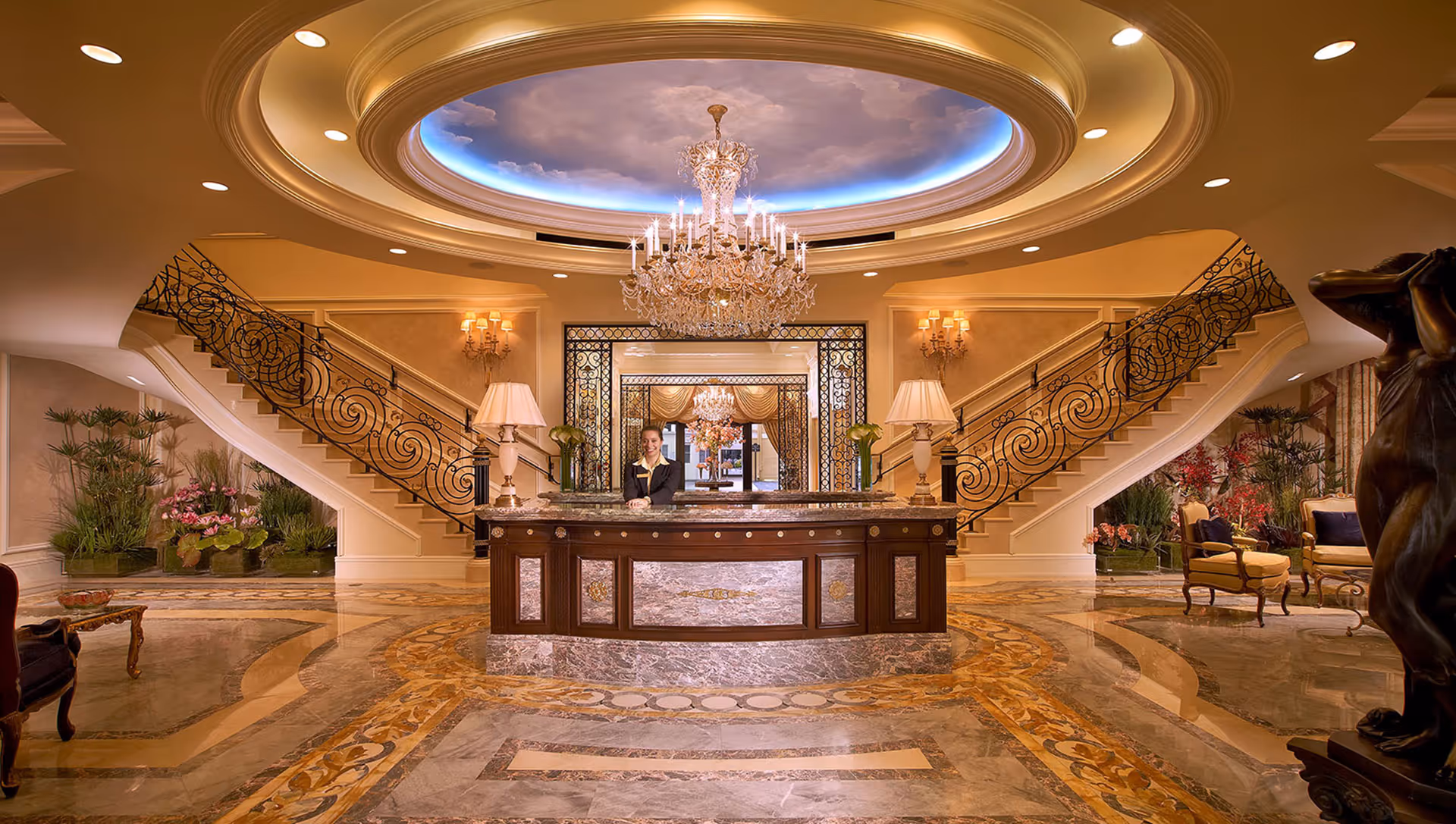 Luxurious lobby area with a central reception desk staffed by a person, ornate double staircases with intricate railings on both sides, elegant chandeliers, warm lighting, and decorative plants and seating areas.