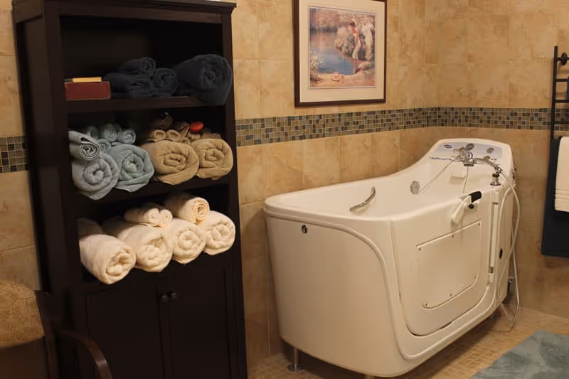 A bathroom with a white walk-in bathtub featuring a door and handrails. Next to the tub is a dark wooden shelving unit filled with neatly rolled towels in various colors including white, beige, light blue, and dark gray. The walls are tiled with beige tiles and a decorative mosaic tile border. A framed picture hangs above the bathtub, and a towel rack with a white towel is visible on the right side.