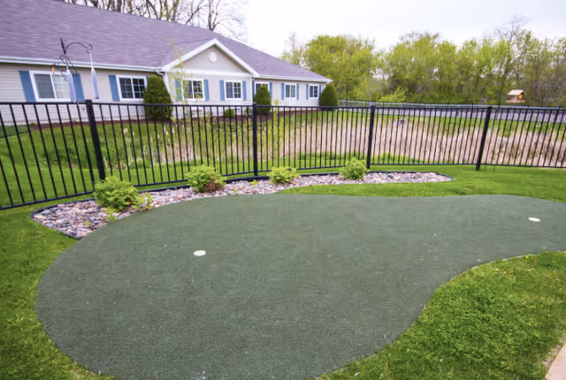 Outdoor putting green area with artificial turf surrounded by grass and a black metal fence. In the background, there is a single-story building with white siding, blue shutters, and a gray roof. Trees and greenery are visible beyond the fence.