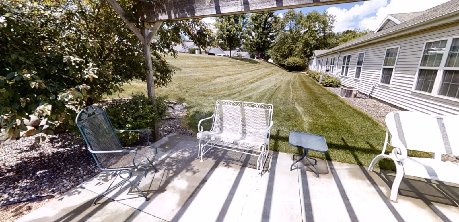 Outdoor patio area with metal chairs and a small table under a pergola, overlooking a well-maintained grassy lawn and a building with multiple windows on the right side.