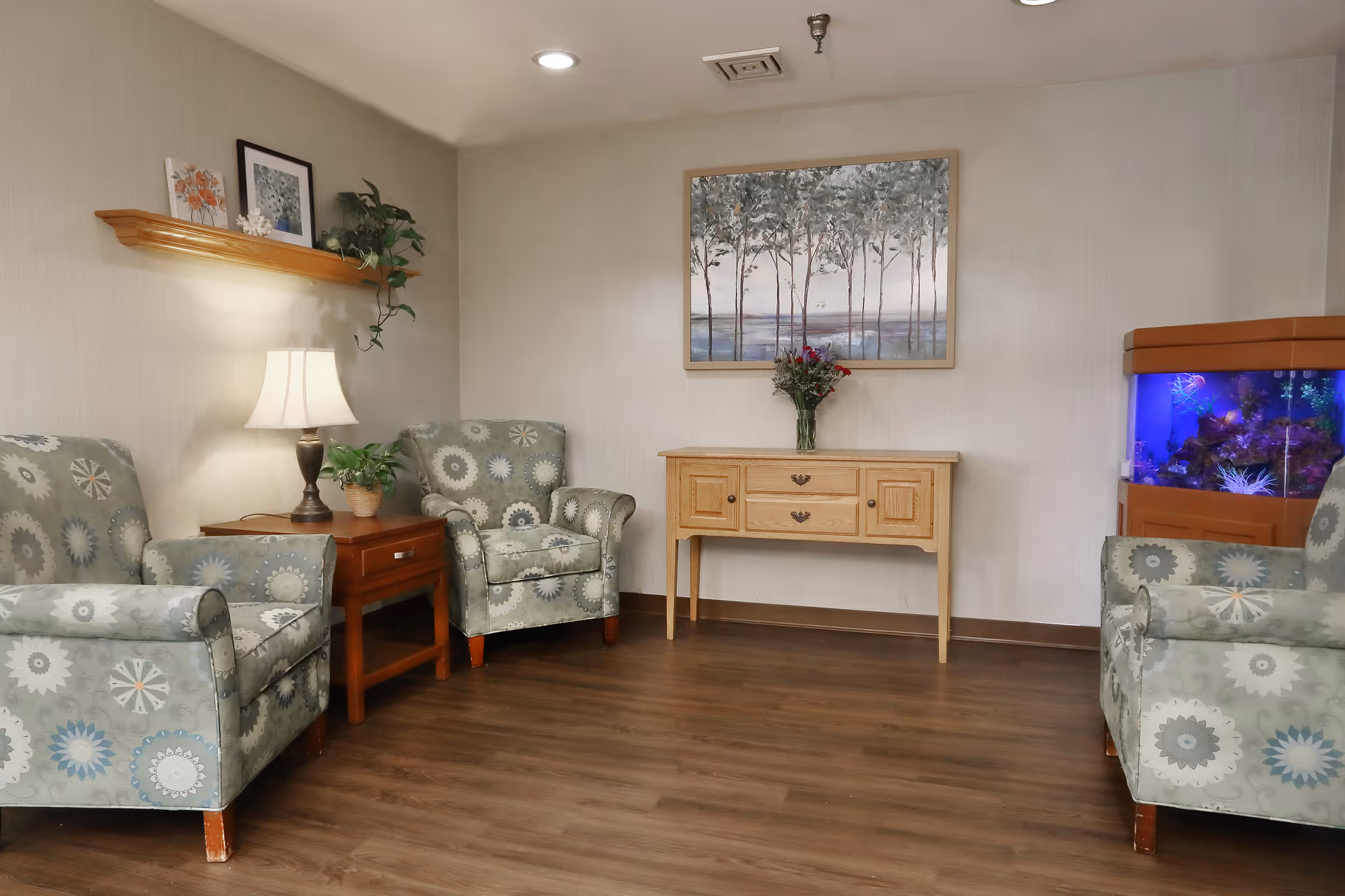 A cozy sitting area with three patterned armchairs arranged around a wooden side table with a lamp and a plant. On the wall above the side table is a wooden shelf holding framed pictures and a plant. Across the room is a wooden console table with a vase of flowers and a large painting of trees above it. To the right is a large fish tank with colorful lighting. The room has wood flooring and neutral-colored walls.