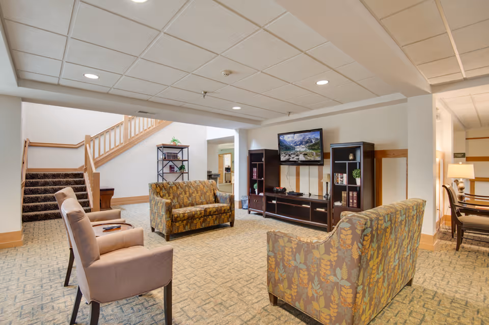 A cozy communal lounge with patterned sofas and chairs facing a wall-mounted TV and shelving, with stairs in the background.