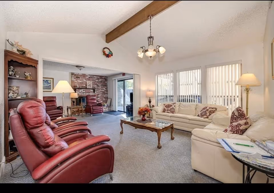 A cozy living room with cream-colored sofas adorned with patterned pillows, two red leather recliners, a wooden coffee table with a flower vase, and multiple lamps providing warm lighting. The room features large windows with vertical blinds, a wooden beam on the ceiling, and an adjoining area with a brick fireplace and additional seating.