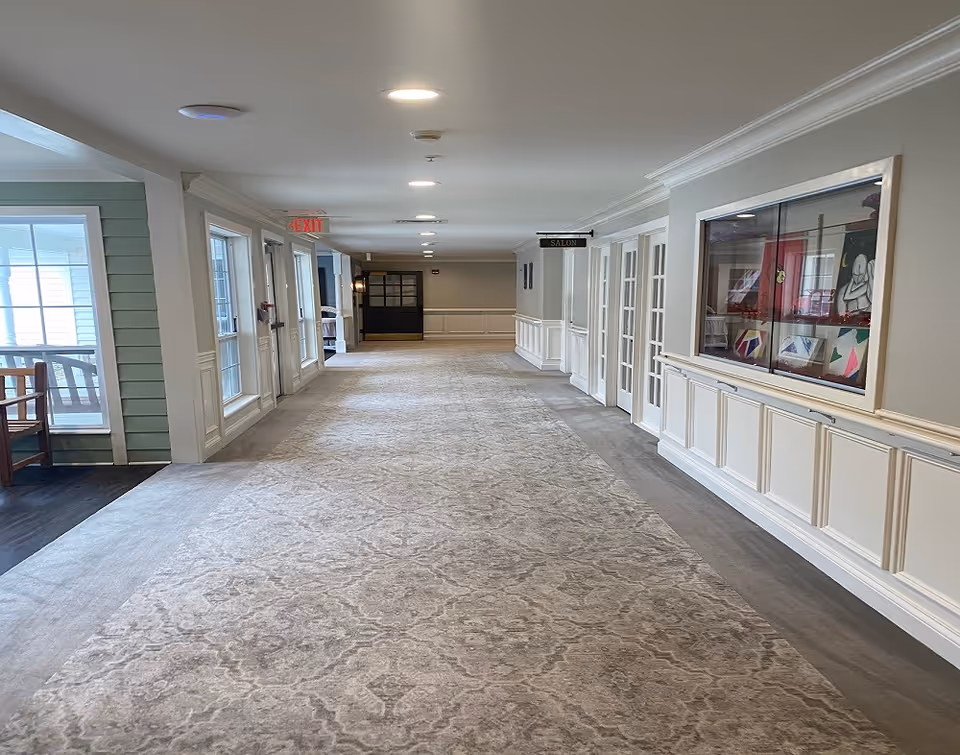 A long, well-lit hallway in a senior living facility with light-colored walls, patterned carpet, and several doors along the right side. There is a display case with artwork on the right wall and a sign indicating a salon. On the left side, there are windows and a seating area with wooden chairs. The hallway has recessed ceiling lights and an exit sign near the far end.