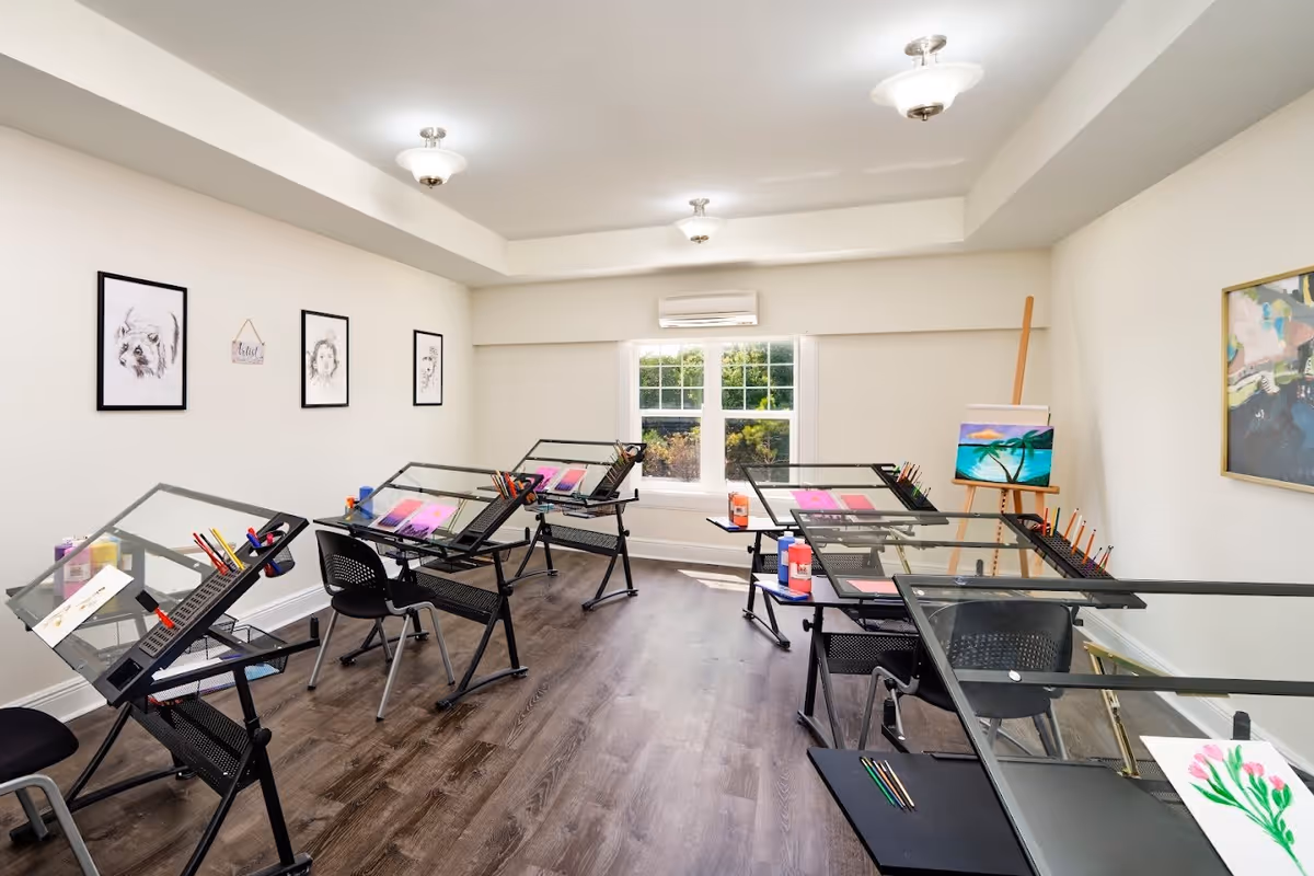 A bright art studio room with six black drafting tables arranged around the space, each equipped with art supplies like colored pencils and paint bottles. The room has white walls adorned with framed sketches and paintings, a large window letting in natural light, and wooden flooring.