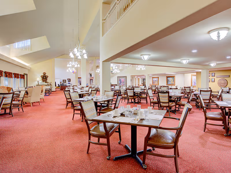 A spacious dining room with multiple tables and chairs arranged neatly on a red carpet. The tables are set with white cups, glasses, and napkins. The room has high ceilings with hanging light fixtures and wall-mounted lights, decorated with framed paintings and a large decorative vase in the background.