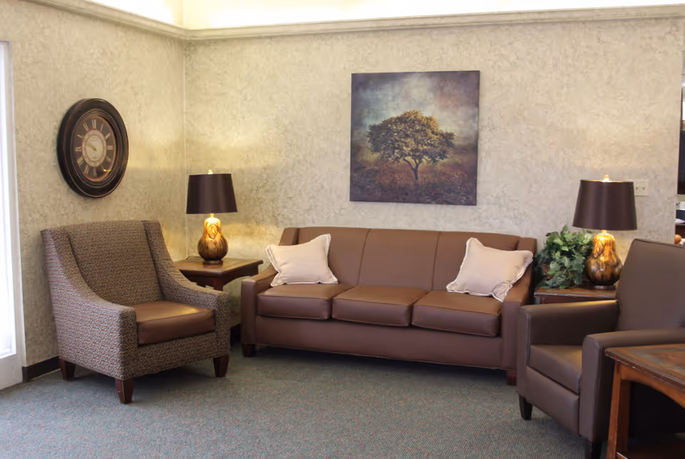 A cozy sitting area with a brown leather sofa adorned with two light-colored pillows, flanked by two armchairs, one patterned and one brown leather. There are two wooden side tables each with a lamp featuring a brown base and dark lampshade. A large wall clock hangs on the left wall, and a framed painting of a tree is centered above the sofa. The walls have a textured light gray finish and the floor is carpeted.