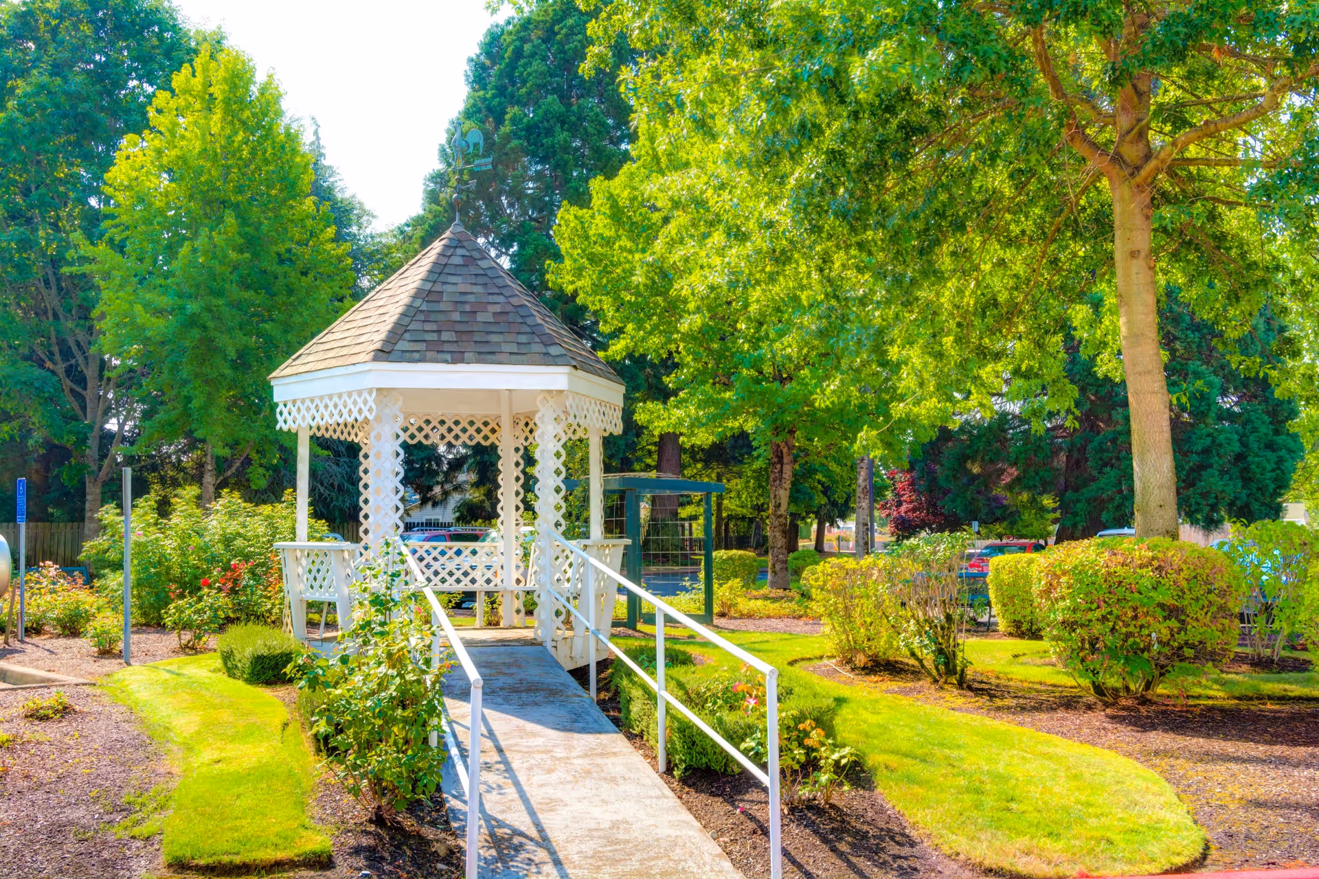 A white wooden gazebo with a shingled roof in a garden area surrounded by green trees, bushes, and manicured grass. A concrete ramp with white railings leads up to the gazebo. The scene is bright and sunny.