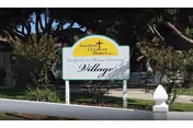 White entrance sign reading 'Sunshine Christian Homes Village' in a landscaped front yard with trees and a white fence post.