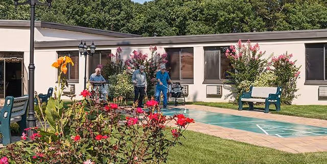 Residents and staff walking and sitting beside a shuffleboard court in a landscaped courtyard with benches, flowering bushes, and a single-story building in the background.