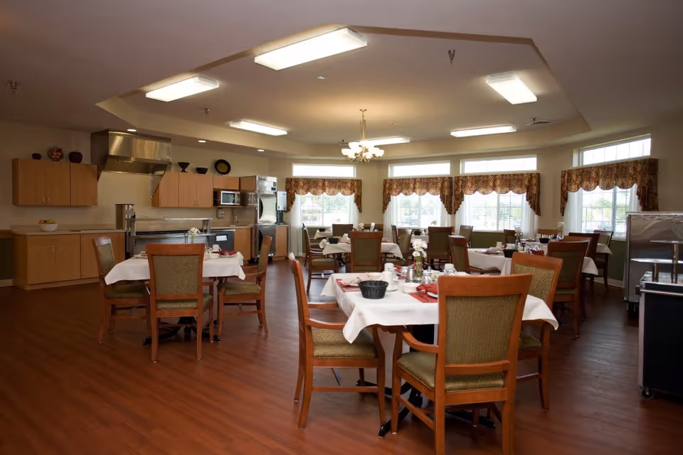 Spacious dining room with multiple set tables and chairs, a serving kitchen in the background, and large windows with valances.