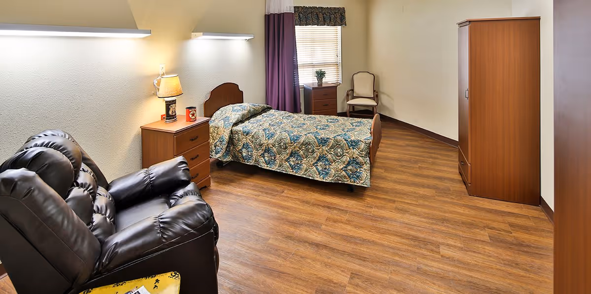 A senior living facility bedroom with a single bed covered in a patterned quilt, a wooden nightstand with a lamp and a small decorative item, a leather recliner chair, a wooden wardrobe, a wooden dresser, a chair near the window with blinds and purple curtains, and wood-look flooring.