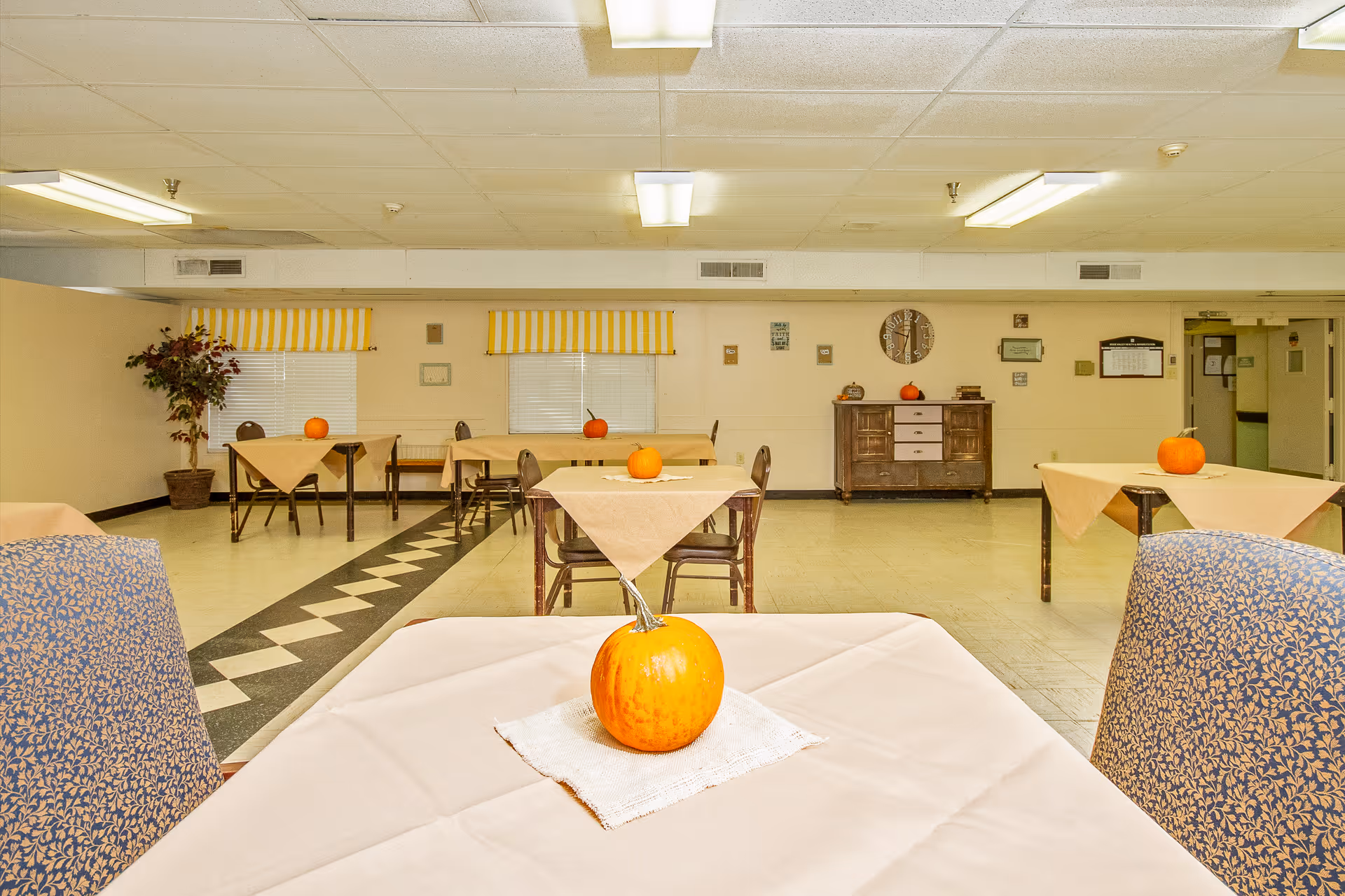 A dining room in a senior living facility with several tables covered in beige tablecloths, each decorated with a small pumpkin. The room has patterned chairs, yellow and white striped window valances, a potted plant in the corner, and a wooden sideboard against the far wall with a clock and additional pumpkins on top.