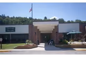 Front entrance of a single-story brick building with a covered walkway, American flag, landscaping and outdoor seating.