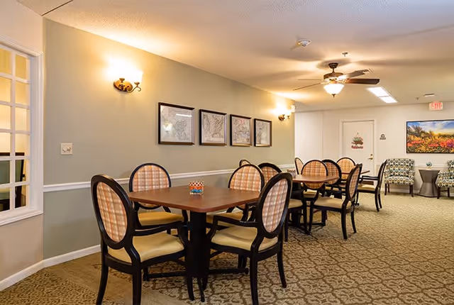 A dining area in a senior living facility with several wooden tables and cushioned chairs arranged neatly. The walls are decorated with framed pictures and wall sconces providing warm lighting. In the background, there are two patterned armchairs with a small table between them and a colorful landscape painting on the wall.