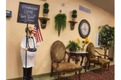 Interior seating area with two ornate chairs and a small table between them, decorated with sunflowers in a vase. A large wall clock and several wall-mounted plants are visible on the beige wall. A statue of a chef holding a chalkboard sign with hours and an American flag stands to the left.