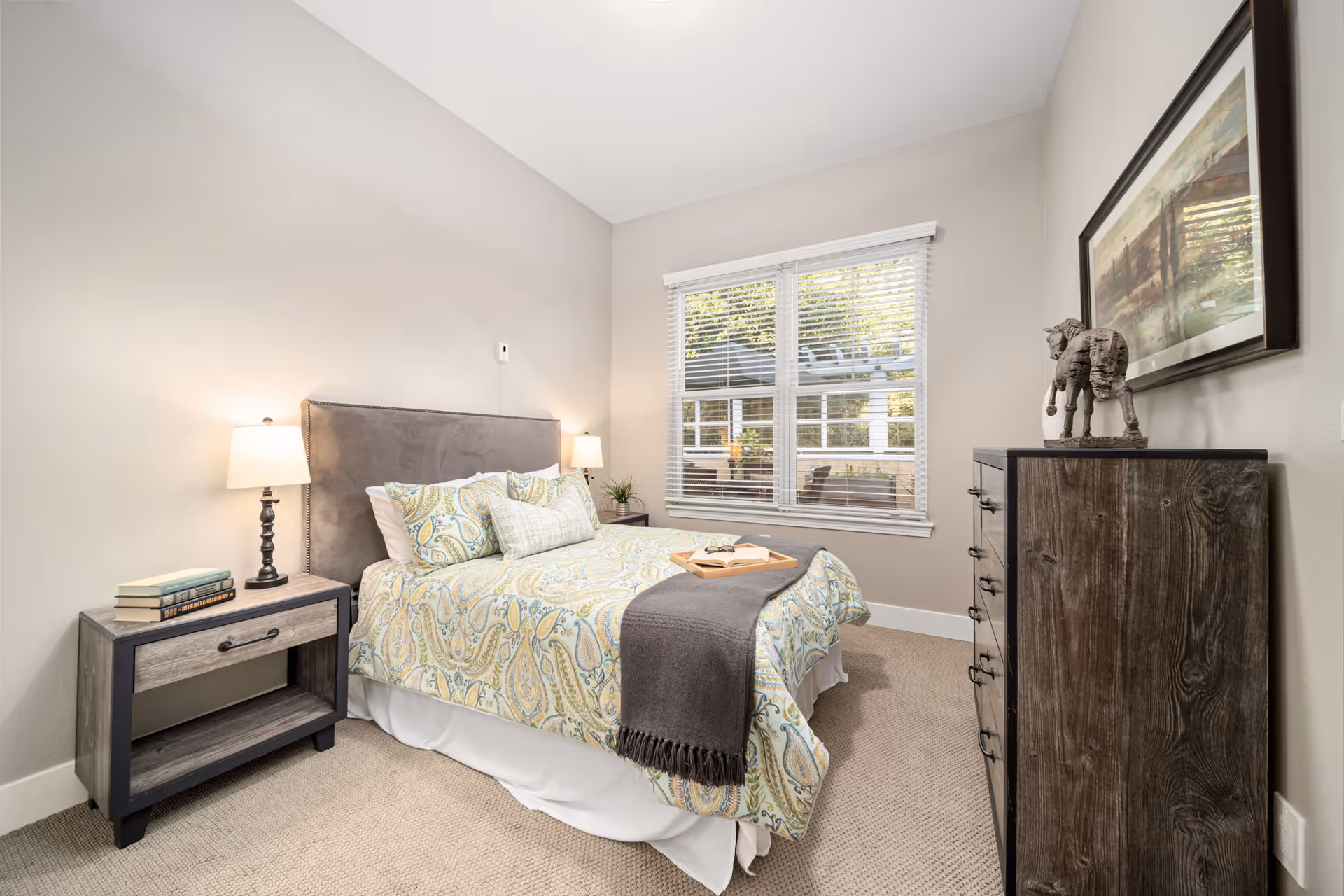 A neatly arranged bedroom with a bed featuring a paisley-patterned comforter and multiple pillows. There is a wooden nightstand with two lamps and a stack of books on the left side of the bed. A wooden dresser with a decorative horse sculpture and a framed painting above it is on the right side. A window with white blinds allows natural light into the room.