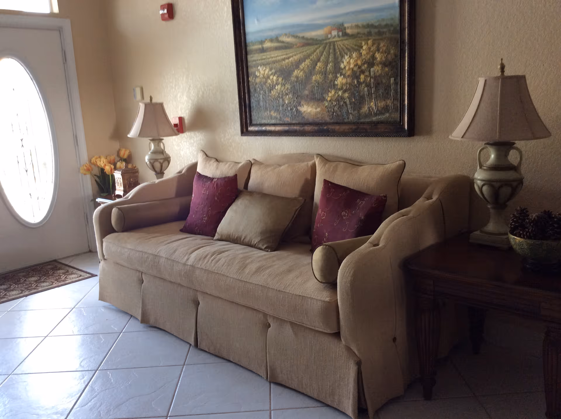 A beige sofa with three cushions, two purple and one beige, placed against a wall with a framed landscape painting above it. There are two side tables on either side of the sofa, each with a decorative lamp. On the left side, there is a door with an oval glass window and a small rug on the tiled floor.