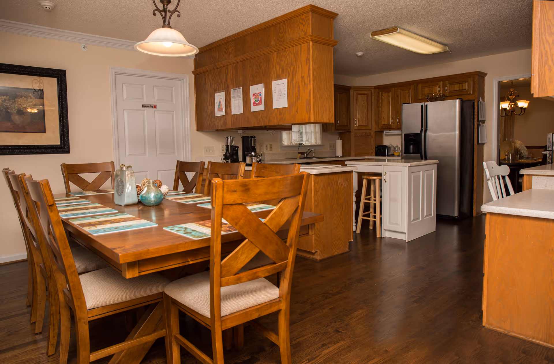 Wooden dining table with place settings and chairs adjacent to a kitchen with an island and stainless steel refrigerator.