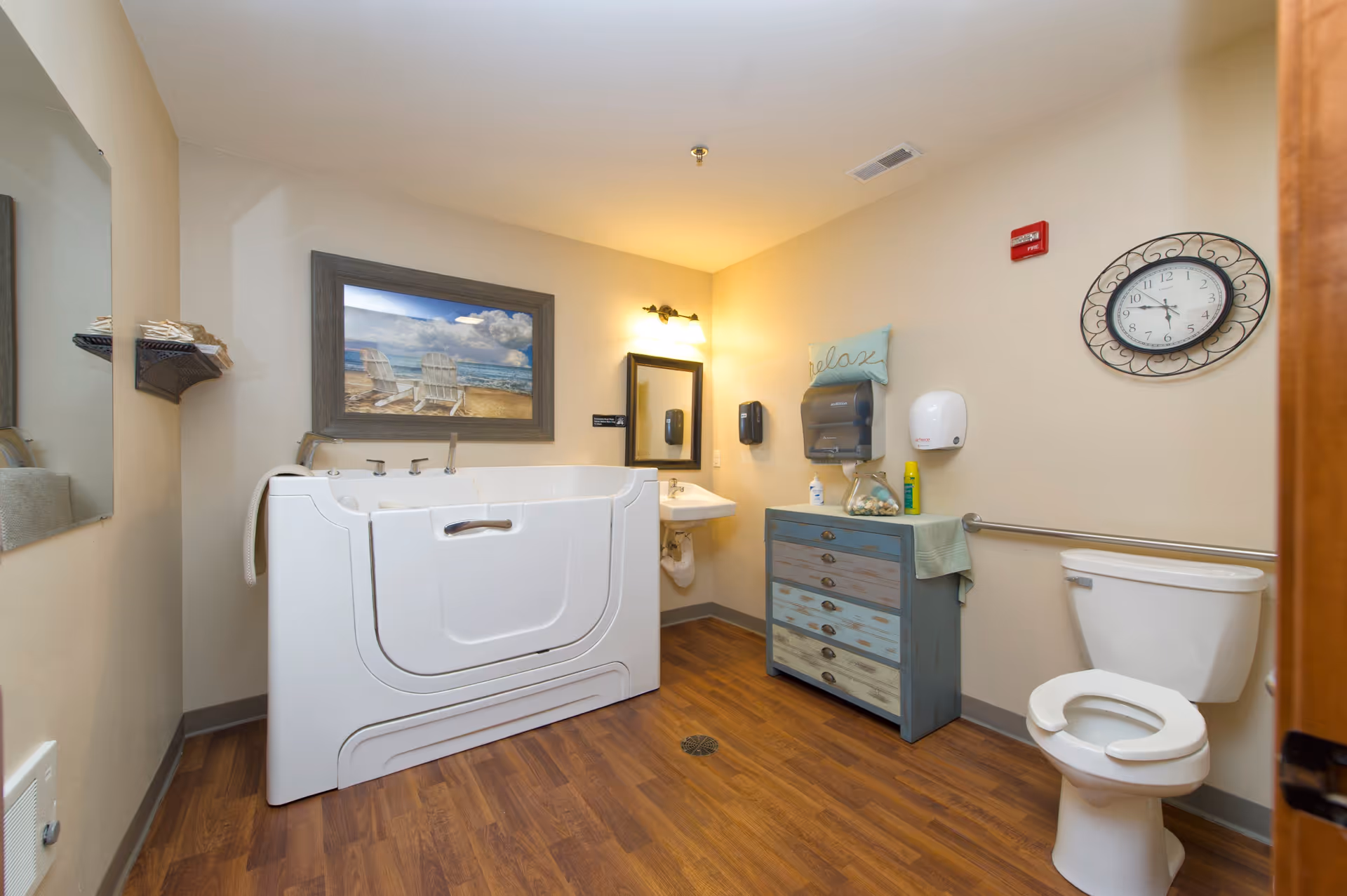A bathroom in a senior living facility featuring a walk-in bathtub with a door, a white toilet with a safety rail, a small sink with a mirror above it, a blue wooden cabinet with drawers, and wall-mounted soap and paper towel dispensers. The walls are beige, and there is a decorative clock and a framed picture of beach chairs on the wall. The floor is wood-patterned.