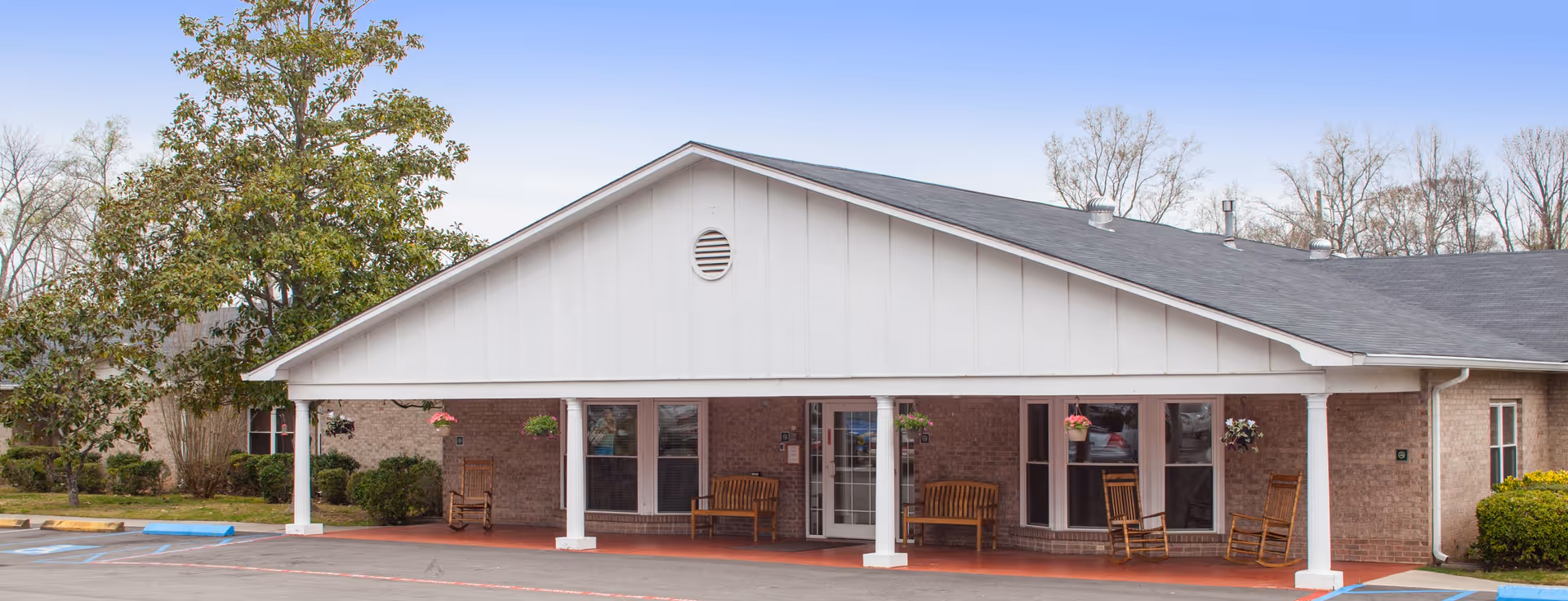 Exterior view of a single-story brick building with a covered porch supported by white columns. The porch has wooden rocking chairs and hanging flower pots. There is a tree and some bushes on the left side, and a parking area in front with marked spaces.