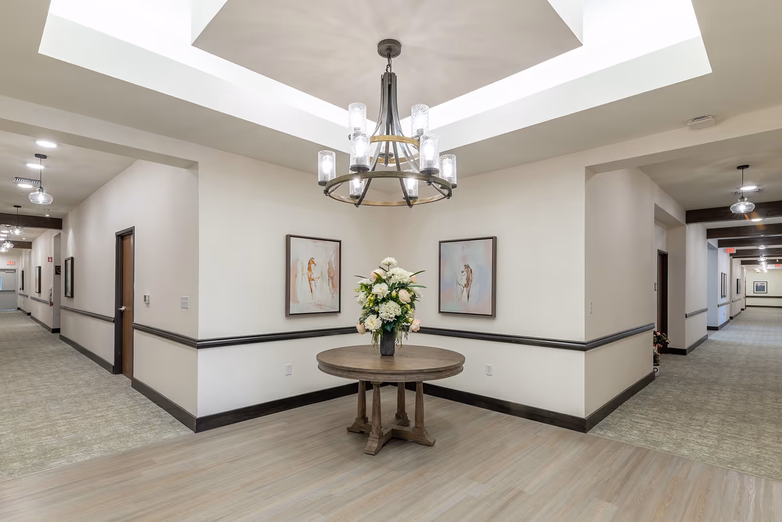 A well-lit hallway intersection in a senior living facility with beige walls and carpeted floors extending in two directions. In the center, there is a round wooden table with a floral arrangement. Above the table hangs a modern chandelier with multiple lights. Two framed paintings of horses are mounted on the walls behind the table.