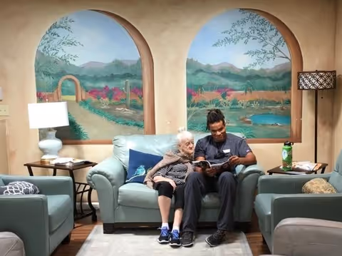 An elderly woman and a caregiver sitting together on a light blue couch in a cozy living room. The caregiver is reading a book to the woman. The room has two large wall murals depicting outdoor desert scenes with mountains, cacti, and colorful flowers. There are two matching armchairs, a floor lamp, and a table lamp on side tables beside the couch.
