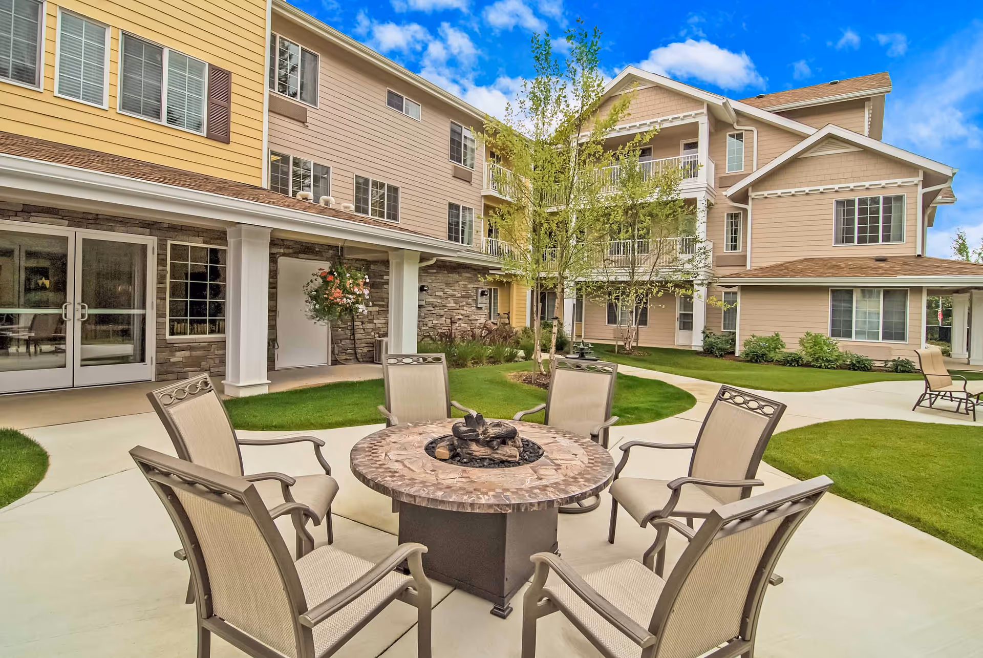 Outdoor courtyard at Holiday Peninsula with a round firepit table surrounded by chairs and a multi-story residential building in the background.
