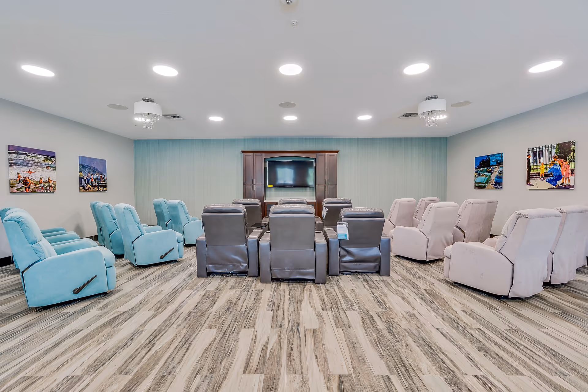 A spacious common room in Senior Point Assisted Living featuring three rows of recliner chairs in blue, gray, and beige colors facing a large flat-screen TV mounted on a wooden cabinet. The room has wood-patterned flooring, light blue and gray walls adorned with colorful paintings, and bright ceiling lights.