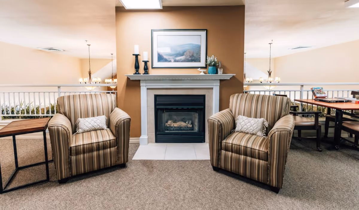 A cozy seating area with two striped armchairs facing a white fireplace with a framed landscape painting above it. There are decorative candles and a small plant on the mantel. To the right, there is a wooden table with chairs, and the area is carpeted with beige walls and ceiling lights.