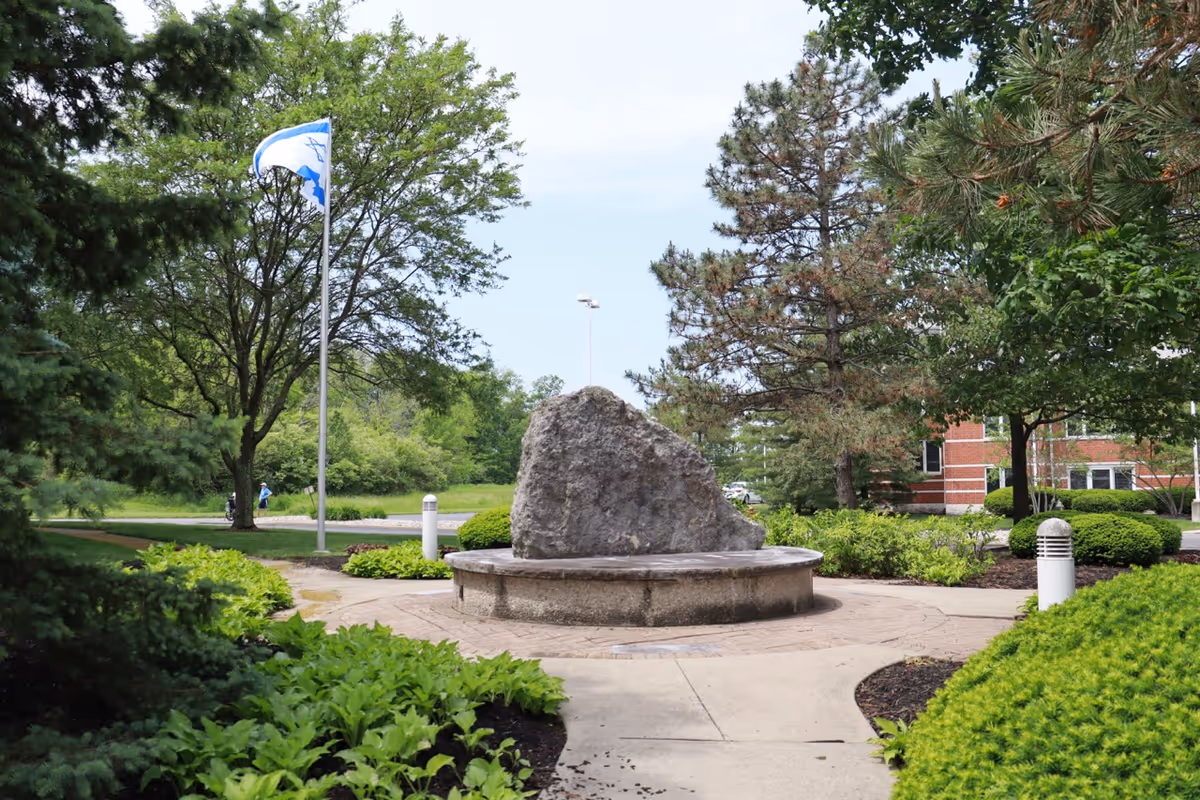 Outdoor garden area with a large rock centerpiece on a circular concrete base, surrounded by green bushes, trees, and a paved walkway. A flagpole with a white and blue flag is visible on the left side, and part of a red brick building is seen in the background on the right.