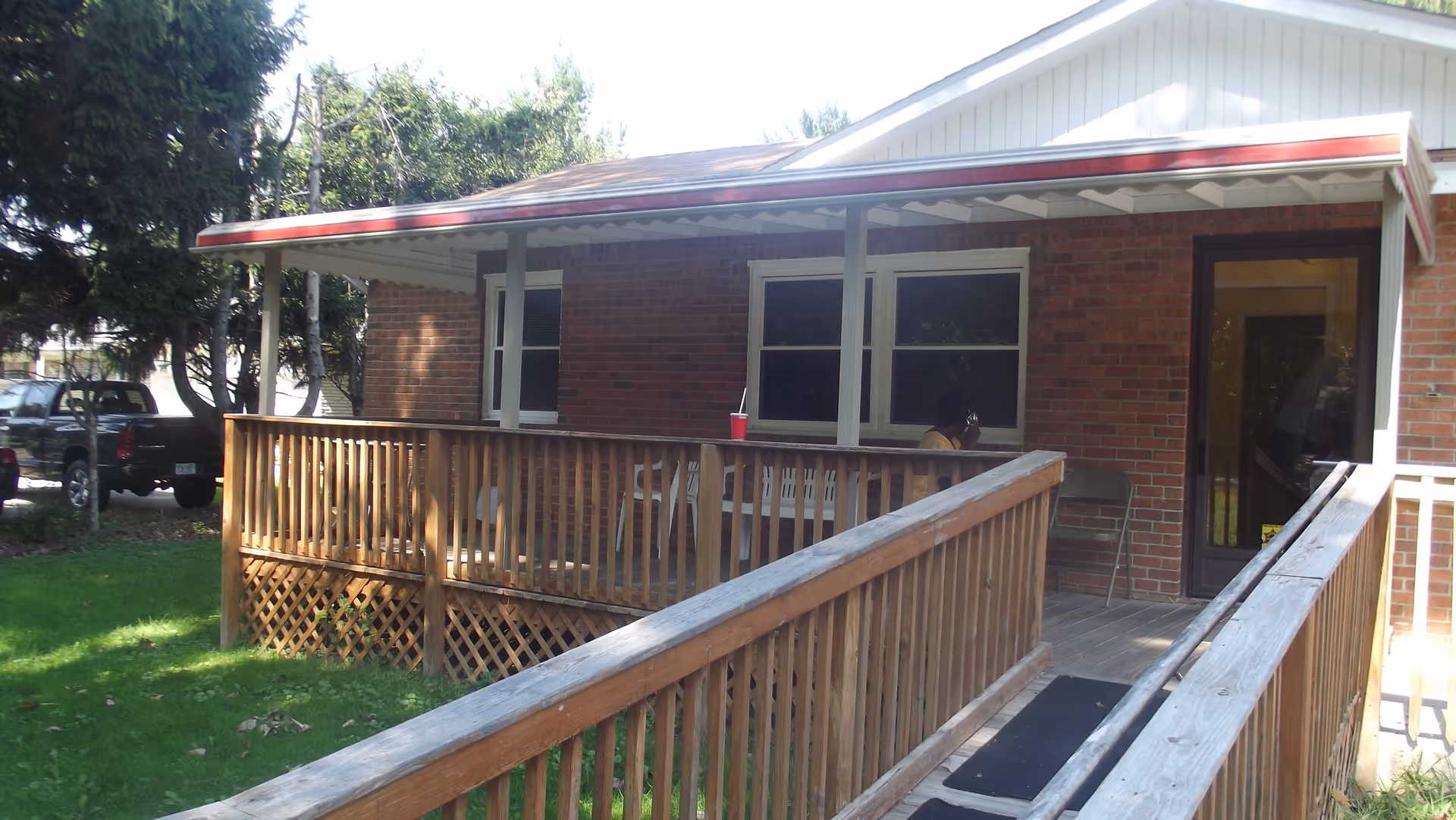 A covered wooden porch with a wheelchair accessible ramp leading to the entrance of a brick building. There is a person sitting on the porch near the windows, and a red cup with a straw is placed on the railing. Trees and parked vehicles are visible in the background.