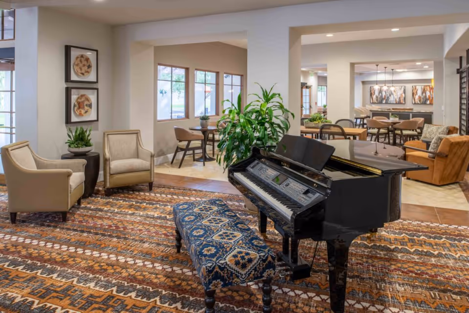 Spacious interior common area featuring a black grand piano with a patterned bench, seating group, plants, and dining tables in the background.