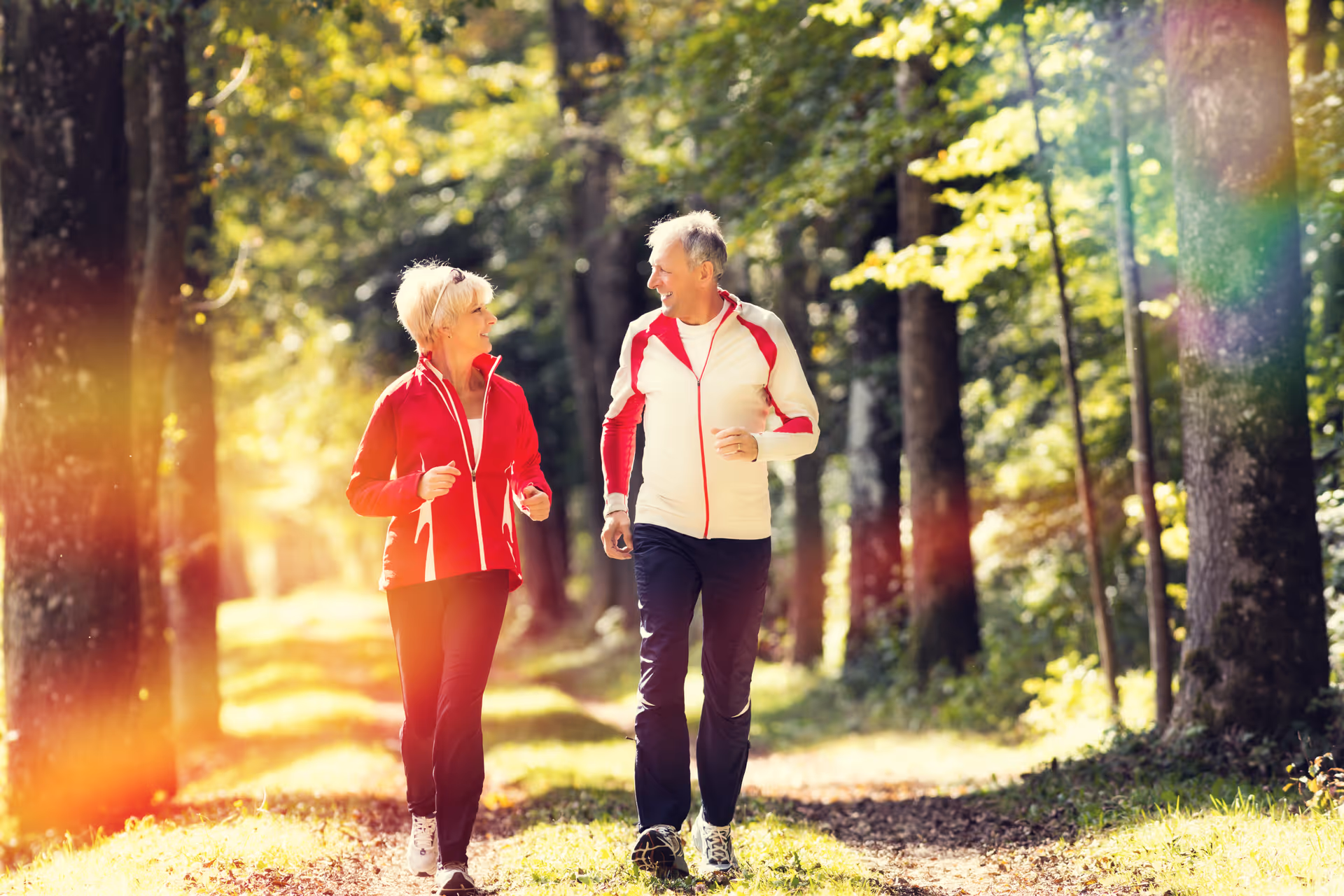 An elderly man and woman jogging together on a forest trail surrounded by tall trees and sunlight filtering through the leaves.