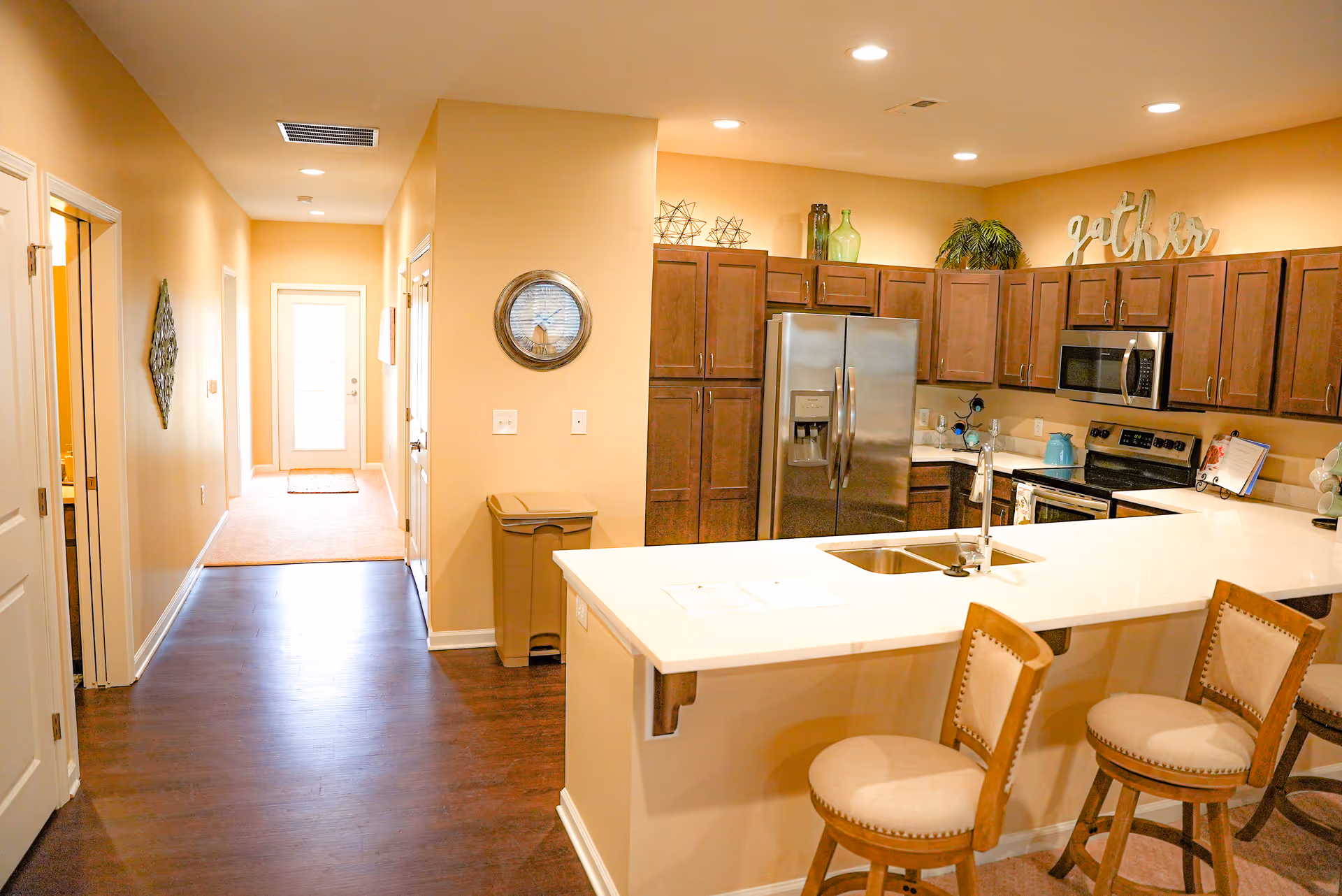Interior view of a kitchen and hallway in a senior living facility. The kitchen features dark wooden cabinets, a stainless steel refrigerator, microwave, and stove, a white countertop with a sink, and three cushioned bar stools. Decorative items and a 'gather' sign are placed on top of the cabinets. The hallway has light-colored walls, dark wood flooring, and leads to a door with frosted glass panels.
