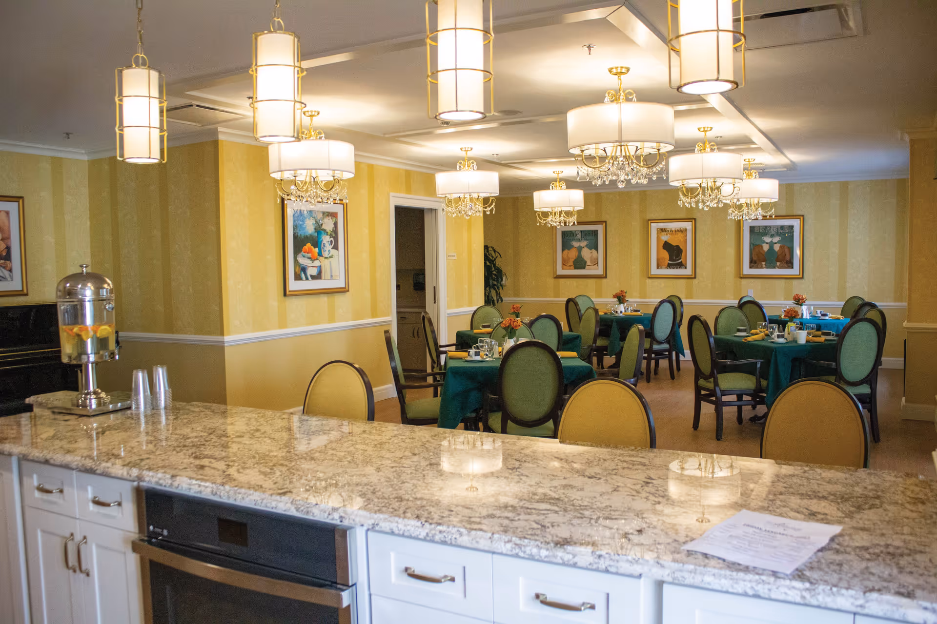 A dining room in a senior living facility with round tables covered in green tablecloths and set with cups, plates, and flowers. The room has yellow striped walls adorned with framed artwork, elegant chandeliers hanging from the ceiling, and a granite countertop in the foreground with a water dispenser and plastic cups.