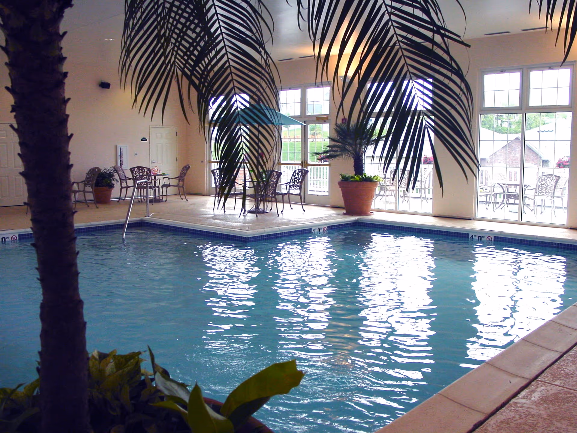 Indoor swimming pool area with clear blue water, surrounded by a tiled deck. There are potted plants including a palm tree and some leafy plants near the pool. In the background, there are large windows and glass doors letting in natural light, with metal chairs and tables arranged near the walls.