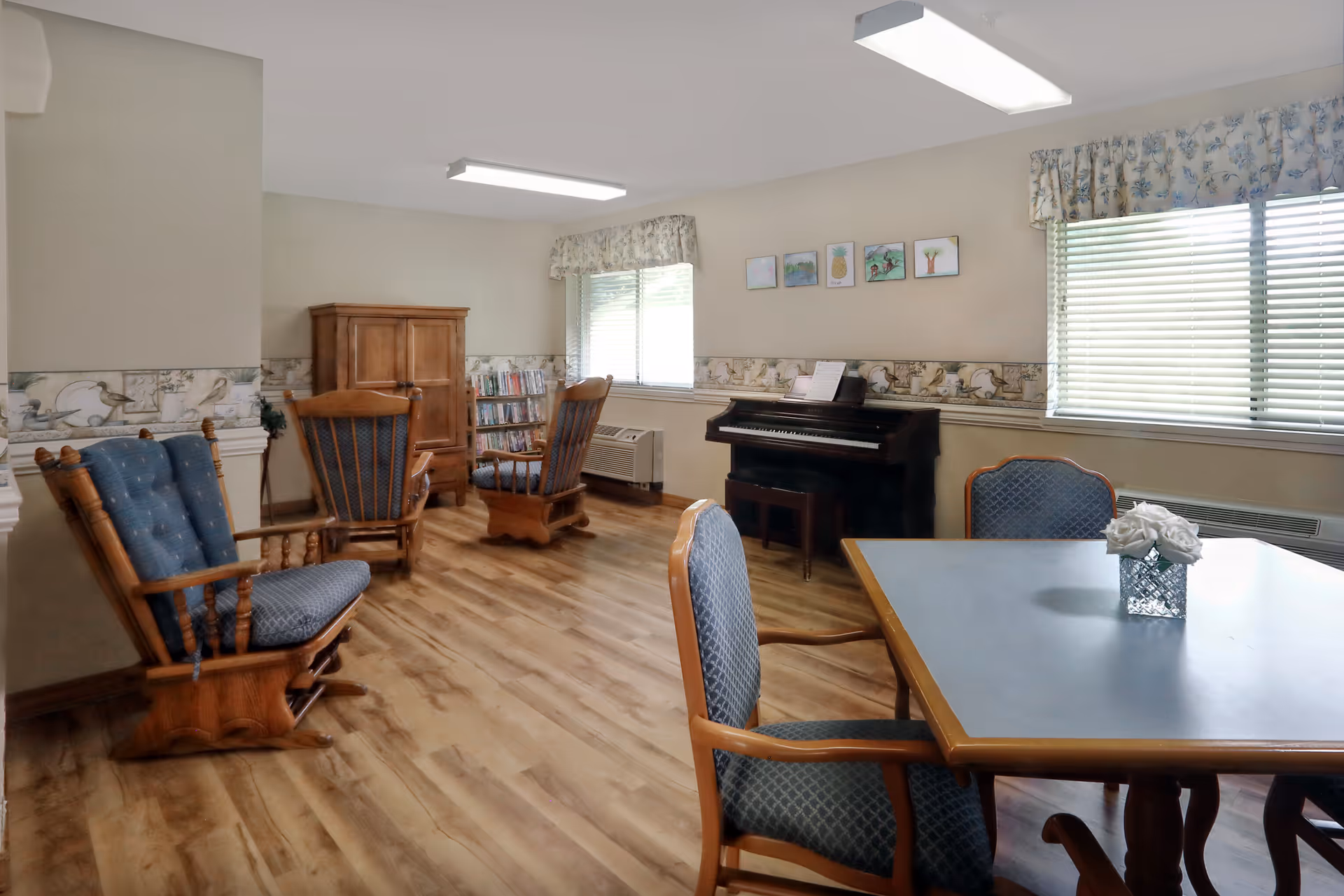 Bright common room with wooden rocking chairs, a table and chairs, and an upright piano by the windows.