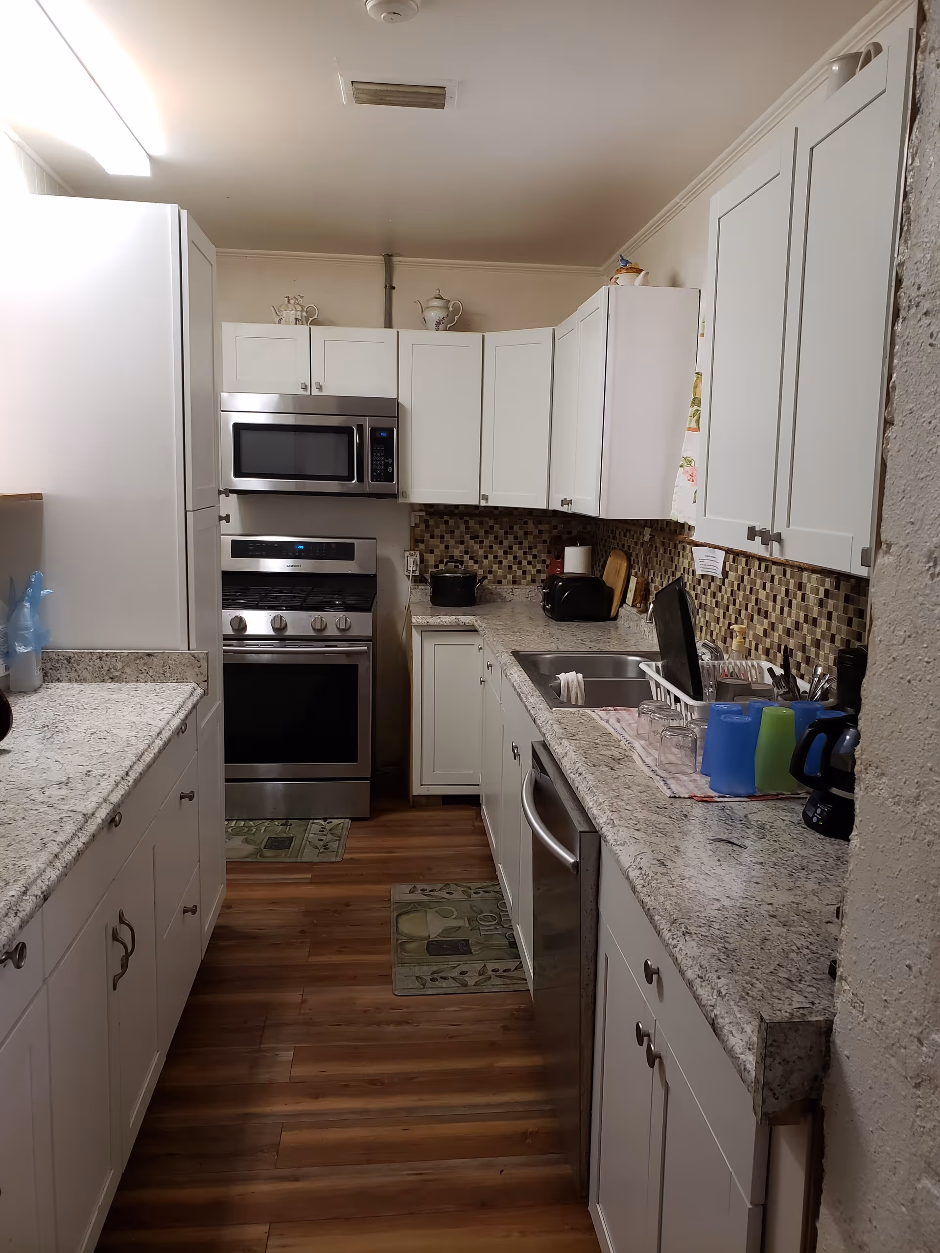 A narrow kitchen with white cabinets, granite countertops, and wood flooring. The kitchen features a stainless steel oven with a microwave above it, a double sink with dishes drying on a rack, a dishwasher, and various kitchen appliances including a toaster and a coffee maker. The backsplash is made of small square tiles in earth tones.