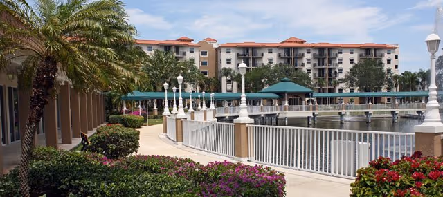 Outdoor view of a senior living facility named Fountainview featuring a paved walkway lined with white lamp posts and railings beside a large pond. There are green bushes, palm trees, and flowering plants along the path. In the background, a multi-story building with balconies and red-tiled roofs is visible under a partly cloudy sky.
