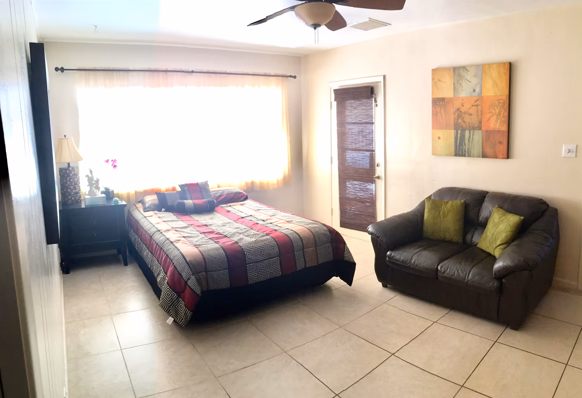 A bright bedroom with a large window covered by sheer curtains. The room features a bed with a patterned bedspread in shades of red, black, and beige, a black nightstand with a lamp and decorative items, a ceiling fan, a dark brown loveseat with two green pillows, and a colorful abstract painting on the wall. There is a door with a bamboo-style blind next to the loveseat.