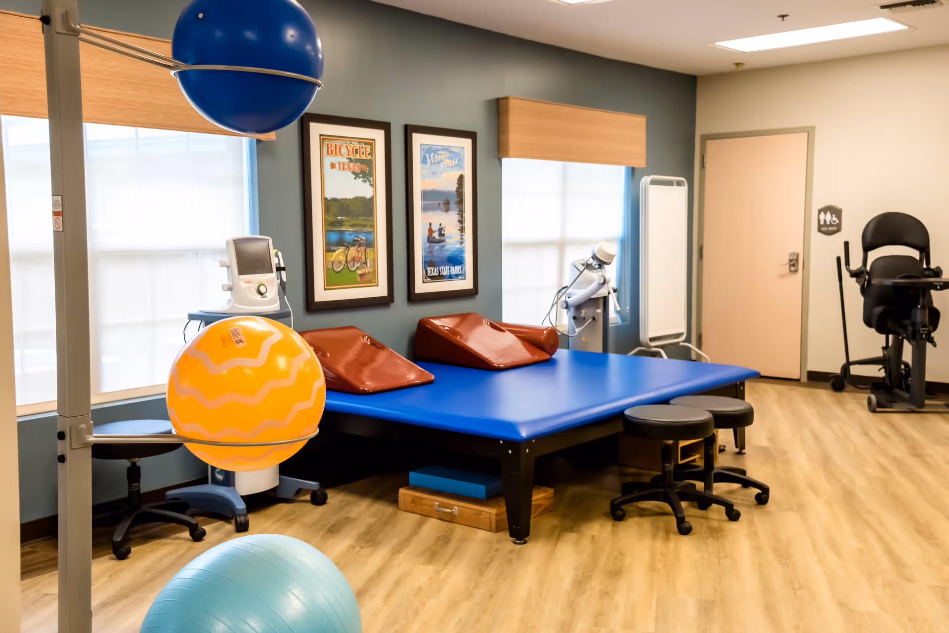 Physical therapy room with a blue therapy table, exercise balls, stools, and exercise equipment. The room has light wood flooring, two framed posters on the wall, and windows with blinds.