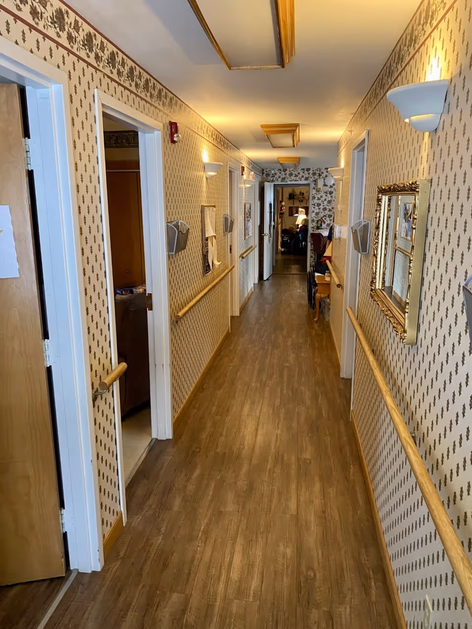 A long, narrow hallway in a senior care facility with wood flooring and patterned wallpaper. The hallway has handrails on both sides, several open doors leading to rooms, wall-mounted lights, and a decorative mirror on the right wall. At the end of the hallway, there is a glimpse of a room with furniture and a lamp.