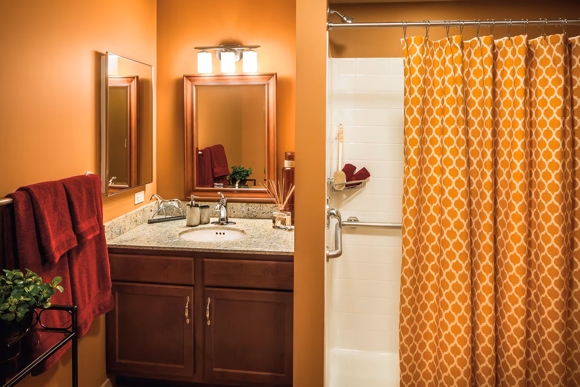 A bathroom with warm orange walls featuring a granite countertop with a sink, a wooden framed mirror above the sink, and a light fixture with three bulbs. There are red towels hanging on a rack and a small green plant on a black metal stand. To the right, there is a shower with white tiles and an orange patterned shower curtain.