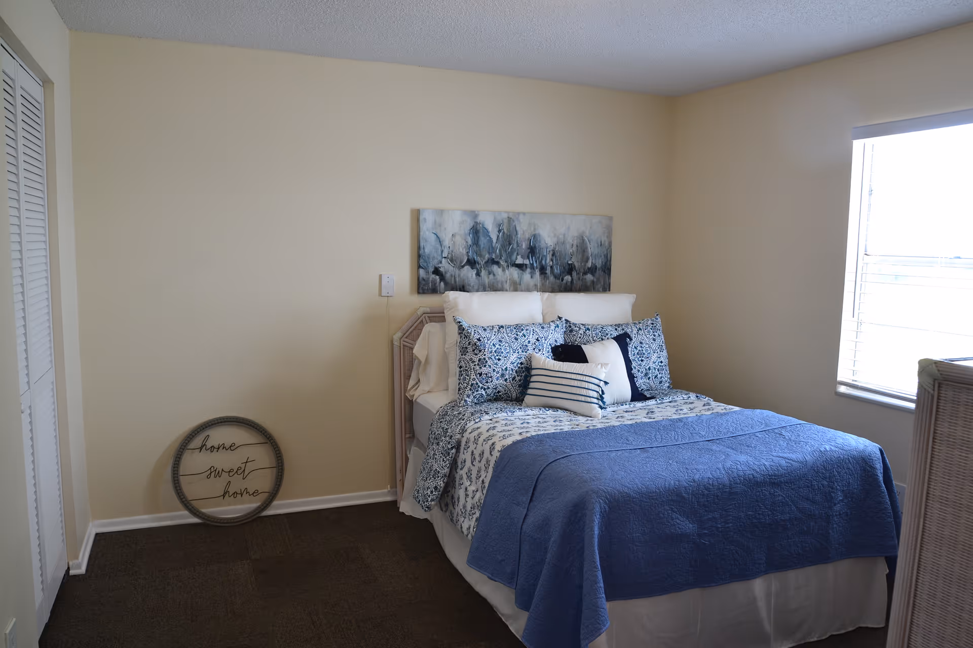 A cozy bedroom with a neatly made bed featuring blue and white bedding and multiple pillows. There is a window with blinds on the right side letting in natural light. A decorative round sign with the words 'home sweet home' leans against the wall near a closed closet with white louvered doors. A piece of abstract artwork hangs above the bed on a beige wall.