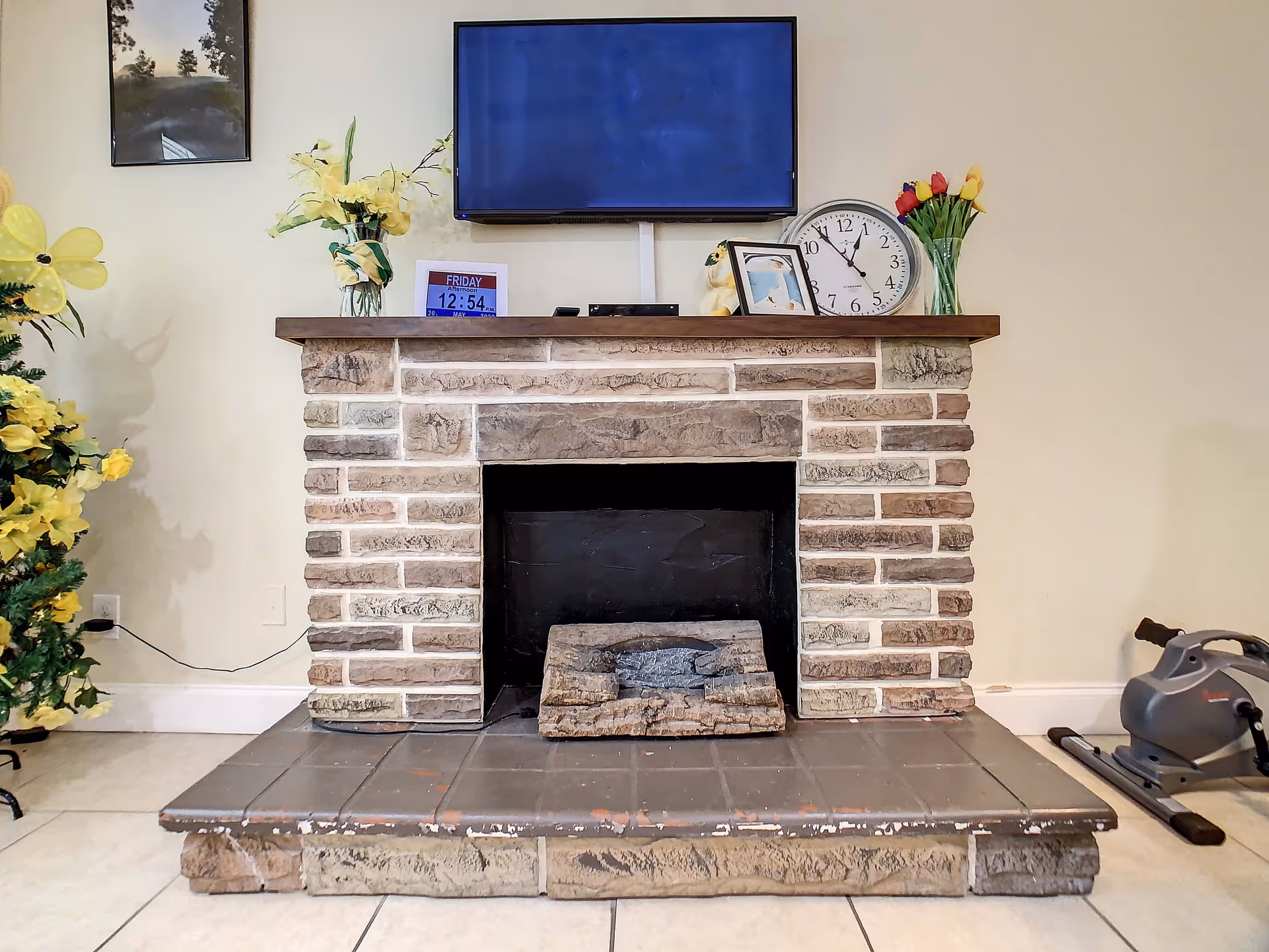 A stone fireplace with a wooden mantel in a living room. On the mantel, there is a flat-screen TV mounted on the wall, a clock, a framed photo, two vases with flowers, and a digital calendar showing the date and time. To the left of the fireplace is a decorated artificial plant with yellow flowers, and to the right is a small exercise machine on the tiled floor.