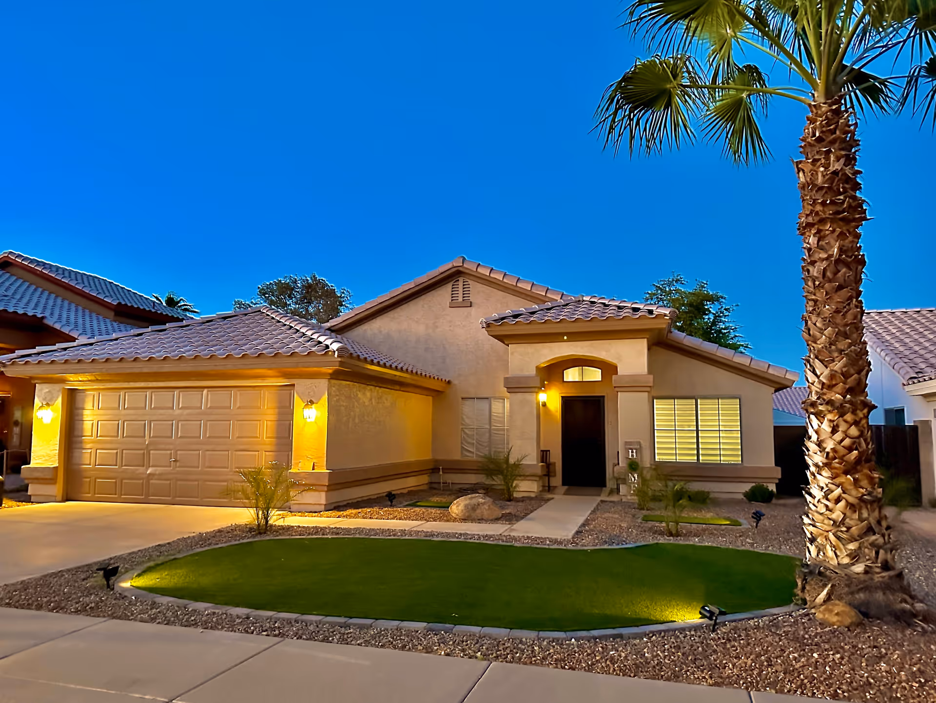 Exterior view of a single-story house at dusk with warm lights illuminating the garage and entrance. The house has a tiled roof, a small front yard with a palm tree, and a well-maintained lawn bordered by rocks.