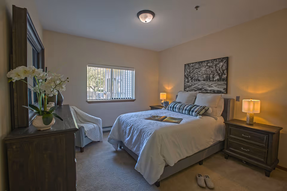 A cozy bedroom with a neatly made bed featuring white bedding and decorative pillows. There are two wooden nightstands on either side of the bed, each with a lit lamp. A large framed black and white picture hangs above the bed. A wooden dresser with a vase of white flowers and a mirror is positioned on the left side of the room. A comfortable chair with a white throw blanket is placed near a window with vertical blinds, allowing natural light to enter. A pair of slippers is on the carpeted floor near the foot of the bed.
