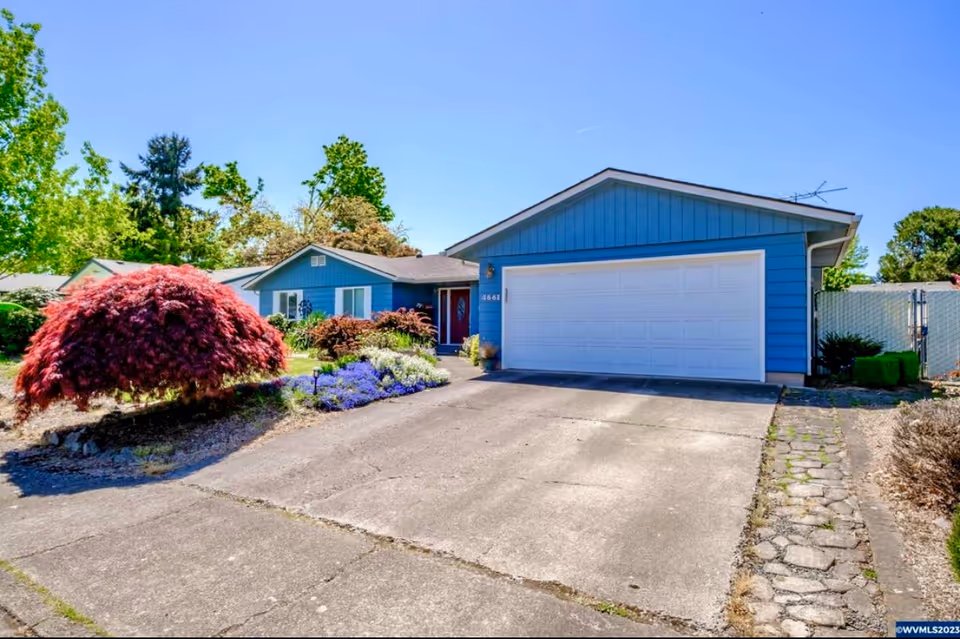 Front exterior view of a single-story blue house with a white garage door, a concrete driveway, and a well-maintained garden featuring a red bush and other greenery under a clear blue sky.